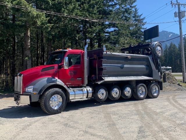 A red DC Materials dump truck on a gravel lot below the mountains
